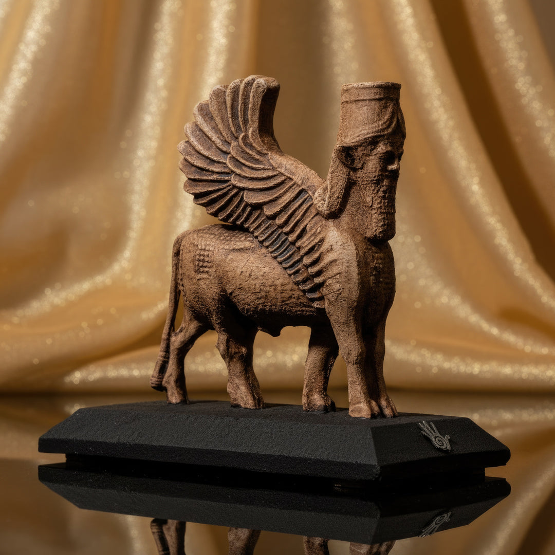 Assyrian Lamassu Statue — ancient Mesopotamian winged bull guardian deity sculpture with human head and divine beard, handcrafted by ArkeoArt. Displayed against golden fabric background highlighting its sacred protective symbolism. Museum-quality archaeological replica, perfect for collectors and historical interior decor.