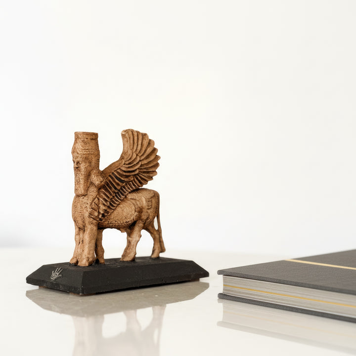 Ahşap masa üzerinde kitap yanında sergilenen Asur Lamassu heykeli – tarih, sanat ve mistik enerjinin birleşimi. / Assyrian Lamassu displayed on wooden desk with books – evokes mystical ancient energy, ideal for libraries, studies, and historical interiors.