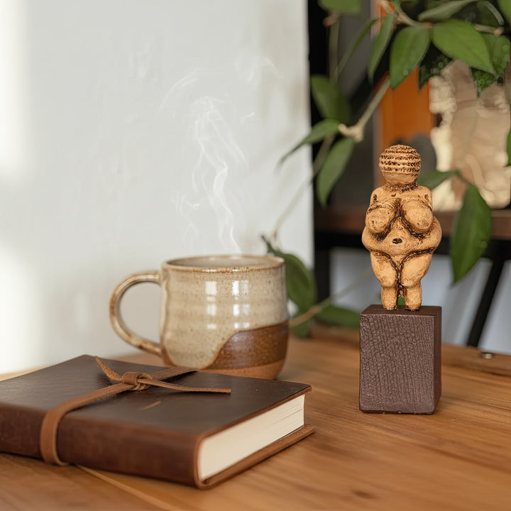 Willendorf Venüsü replikası kitap ve kahve kupası ile birlikte masa üzerinde sergilenen lifestyle fotoğrafı. Paleolitik dönemin ünlü Venus of Willendorf figürü, modern iç mekânda dekoratif arkeoloji objesi olarak görülmektedir. ArkeoArt tarafından üretilen bu Paleolithic fertility goddess sculpture, tarih öncesi sanat ve arkeoloji koleksiyonları için güçlü bir dekoratif sanat eseridir.